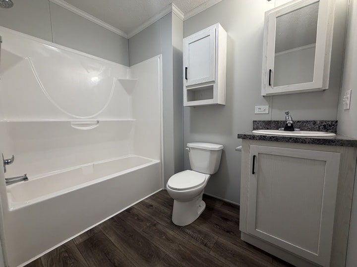 A clean, modern bathroom with a white bathtub and shower combo, a toilet, a vanity with a sink, medicine cabinet, wall-mounted cabinet, and wood-style flooring. Walls are painted light gray.