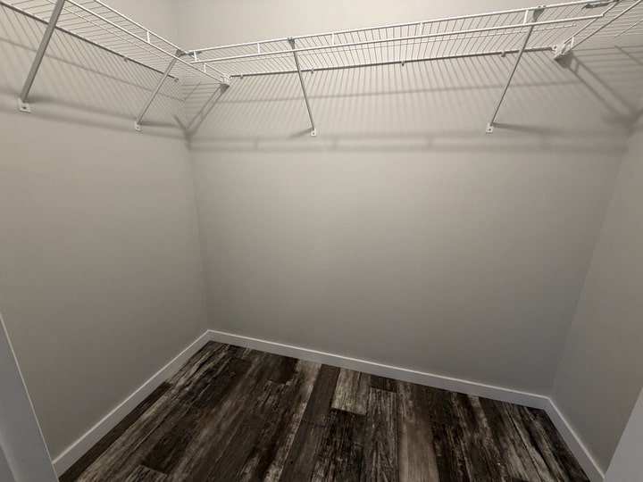 Empty walk-in closet with white walls, wire shelves along two walls, and dark wood-patterned flooring. The closet is well-lit and has a clean, modern appearance.