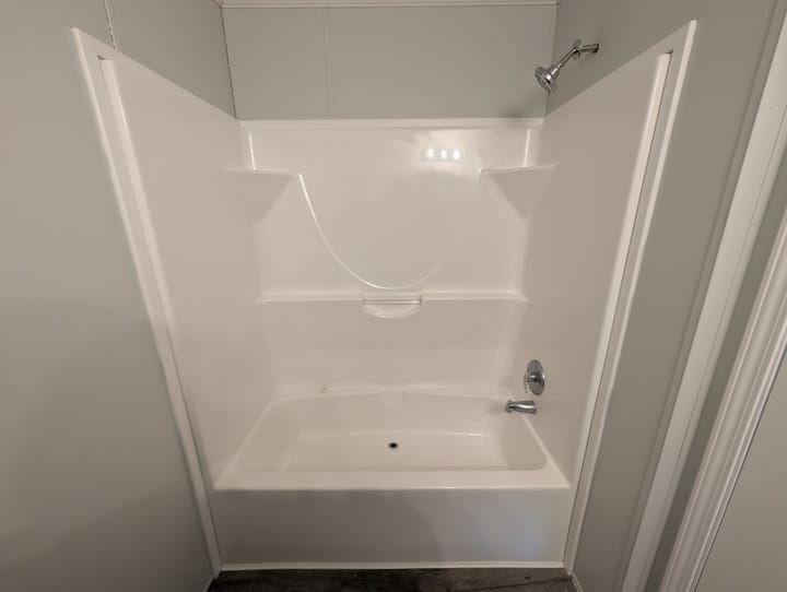 A white bathtub and shower combo with a built-in curved back wall, shelves, faucet, and showerhead, set against light gray walls in a bathroom.