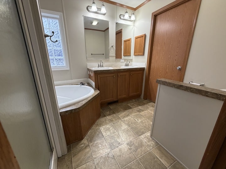 Bathroom with a large corner bathtub, double sinks with mirrors and lights above, wood cabinets and doors, a glass shower door, and tiled floor. A small partition separates the toilet area.