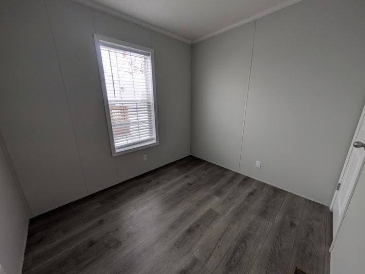 Small empty room with light gray walls, a single window with white blinds, dark wood-style flooring, and a white door partially visible on the right. Natural light enters through the window.