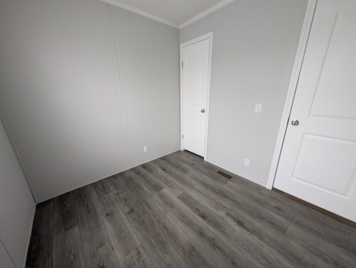 A small, empty room with light gray walls, white trim, two white doors, and dark gray wood-look flooring. The space is well-lit and has a modern, minimal appearance.