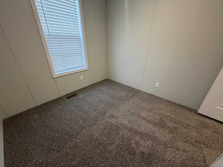 A small, empty room with light gray walls, a window with closed white blinds, and carpeted flooring in a neutral gray shade. There are wall outlets and a door partially visible on the right.
