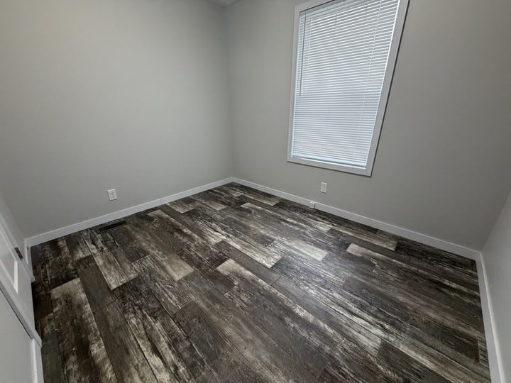 Empty room with light gray walls, a single large window with closed blinds, dark wood-style flooring, and white trim. The space is well-lit and minimalist, with no furniture or decorations.