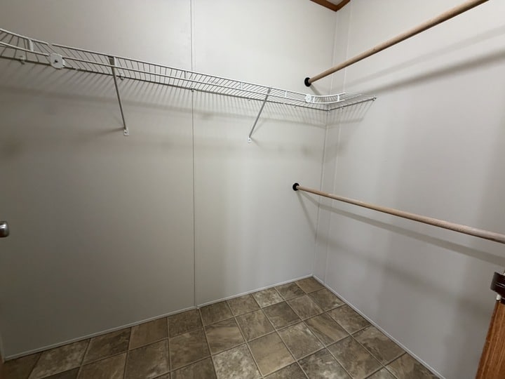 Empty walk-in closet with beige tile flooring, white walls, a metal wire shelf with hang rod on the left, and two wooden closet rods on the right wall.