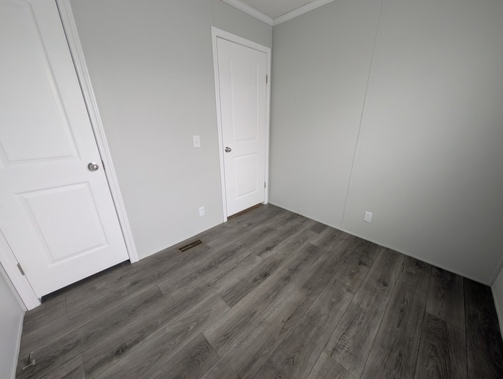A small empty room with light gray walls, two white doors, white trim, and gray wood-look flooring. There is a floor vent and an electrical outlet visible.