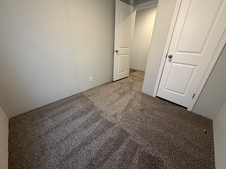Small empty room with light gray walls, clean brown carpet, and two white doors—one open and one closed. The room is lit by natural or overhead light.