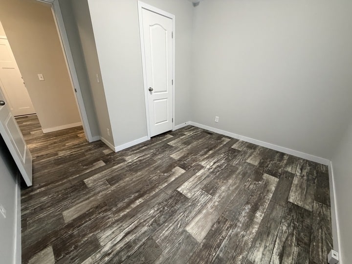 A small empty room with grayish wood-patterned flooring, light gray walls, a white door, and baseboards. The room is well-lit and clean, with an open doorway leading to another area.