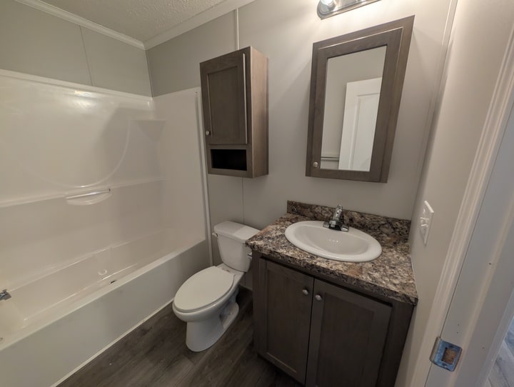 A small bathroom with a bathtub and shower, a toilet, a sink with a granite-style counter, a mirrored medicine cabinet, and a wall-mounted storage cabinet, all in neutral tones.