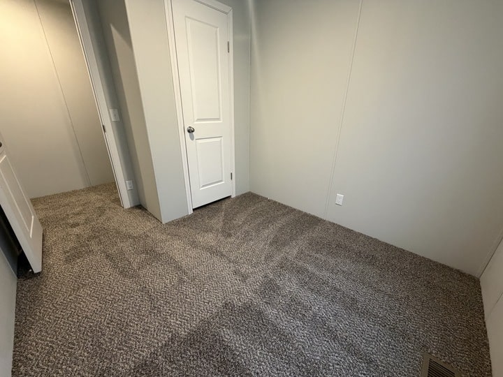 A small, empty room with beige carpet, light gray walls, a white door, and a vent on the floor. The room is well-lit, and the carpet shows vacuum lines.