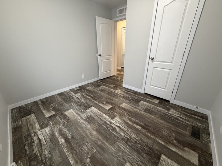 A small, empty room with gray wood-patterned flooring, light gray walls, and two white doors, one slightly open. The room appears clean and well-lit with no furniture.