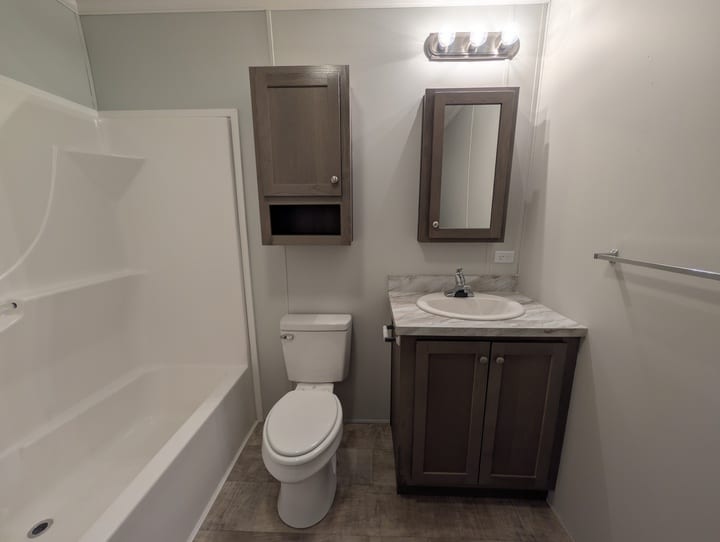 A small bathroom with a bathtub and shower on the left, a toilet in the center, and a sink with a marble countertop, cabinets, and a mirrored medicine cabinet on the right. Walls are light-colored.