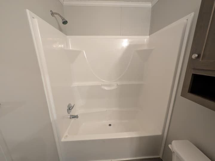 White bathtub and shower combination with built-in shelves, silver faucet, and showerhead in a clean, modern bathroom with light gray walls. Part of a toilet and wooden cabinet are visible on the right.