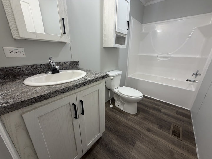A modern bathroom with gray walls, white cabinets, a granite countertop with sink, a toilet, and a white bathtub-shower combo. The floor is dark wood-style vinyl.