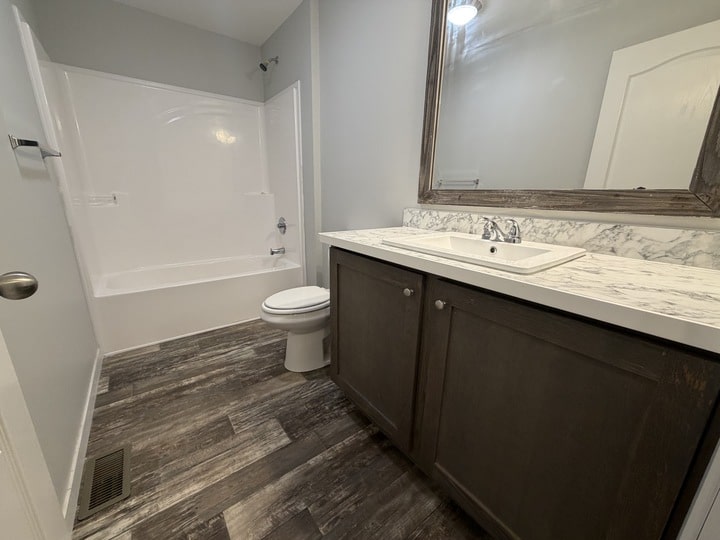 A modern bathroom with gray wood-look flooring, a white bathtub and toilet, and a vanity with a marble countertop and dark wood cabinets. A large framed mirror hangs above the sink.