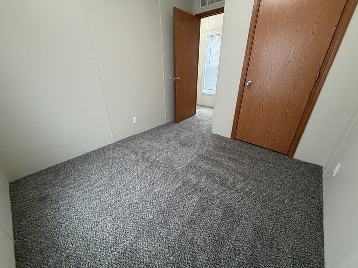 An empty room with light-colored walls, gray carpet, and two wooden doors—one open, leading to another room with a window, and the other closed. The room appears clean and undecorated.