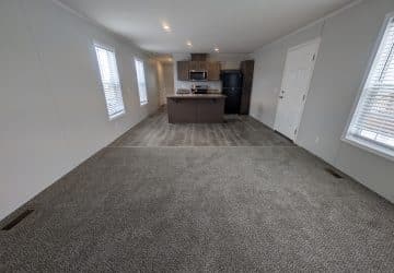 Open floor plan with carpeted living area, transitioning to a kitchen with wood-style flooring, brown cabinets, black appliances, and three large windows letting in natural light.