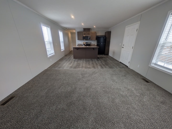 Open floor plan with carpeted living area, transitioning to a kitchen with wood-style flooring, brown cabinets, black appliances, and three large windows letting in natural light.