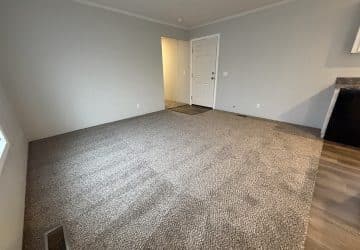 A mostly empty room with light gray carpet, pale gray walls, a white door, and a small section of wood flooring near the entrance. There is a vent on the carpet and a countertop partially visible on the right.