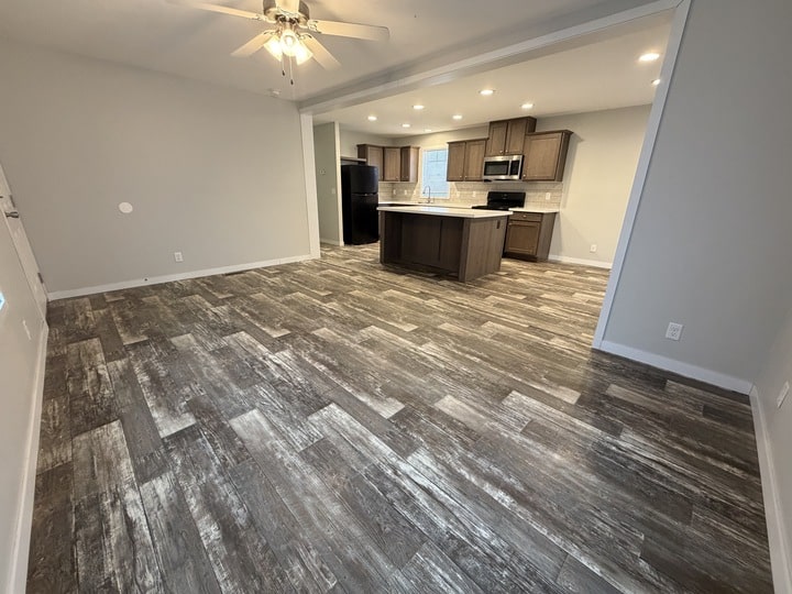 Open concept living area with gray wood-look tile flooring, beige walls, a ceiling fan, and a modern kitchen featuring dark wood cabinets, an island, black appliances, and recessed lighting.