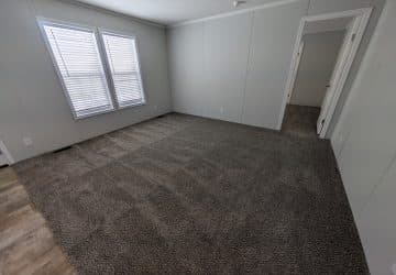 Empty room with gray carpet, light gray walls, two large windows with blinds, and a doorway leading to another carpeted room. The edge of a laminate wood floor is visible in the foreground.