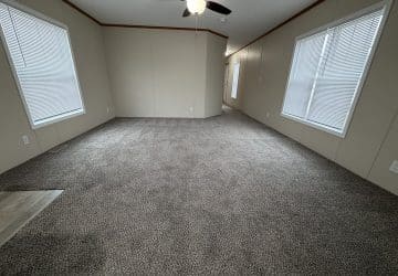A spacious, empty room with beige walls, gray carpet, two large windows with blinds, and a ceiling fan with lights. The room has no furniture and is well-lit by natural light from the windows.