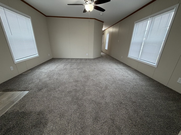A spacious, empty room with beige walls, gray carpet, two large windows with blinds, and a ceiling fan with lights. The room has no furniture and is well-lit by natural light from the windows.