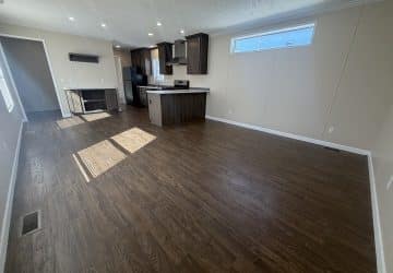 A spacious, unfurnished room with dark wood flooring, beige walls, and a kitchen area featuring dark cabinets, an island, and stainless steel appliances. Sunlight streams through windows, creating bright patches on the floor.