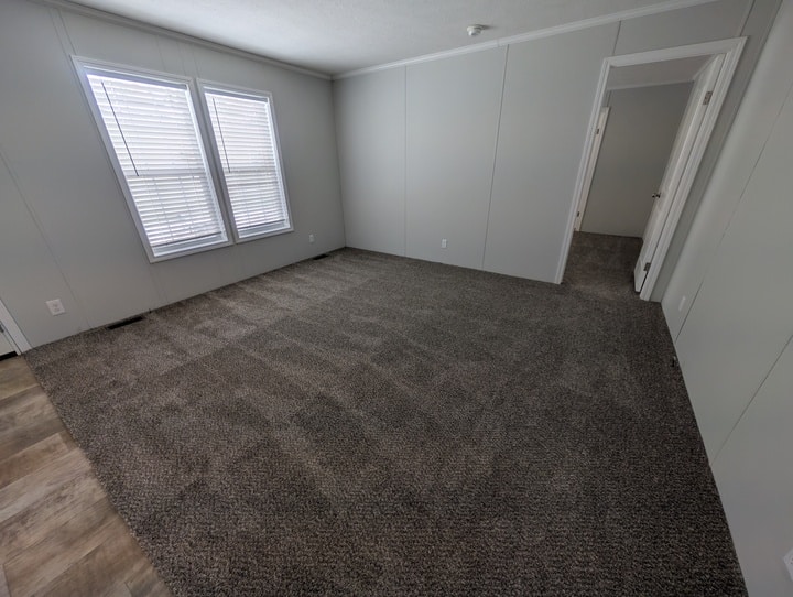 Empty room with gray carpet, light gray walls, two large windows with blinds, and a doorway leading to another carpeted room. The edge of a laminate wood floor is visible in the foreground.