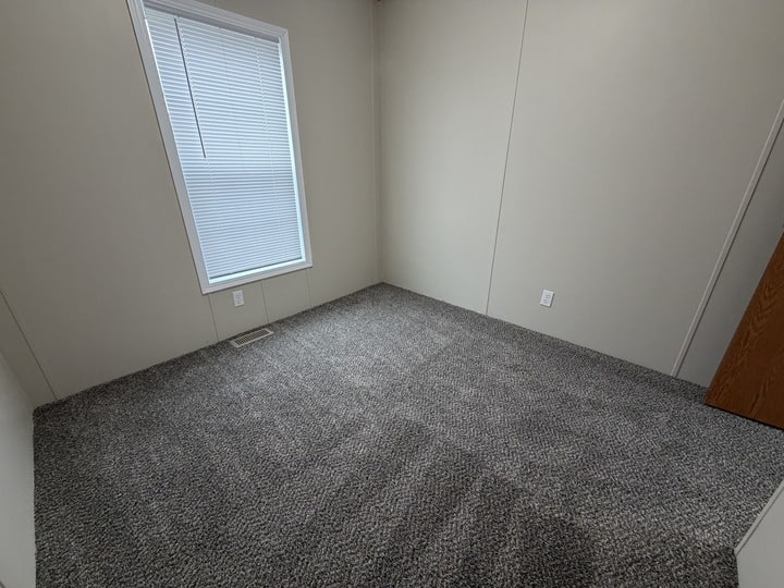 An empty room with light beige walls, a closed window with blinds, gray carpet flooring, and an open wooden door visible on the right side.