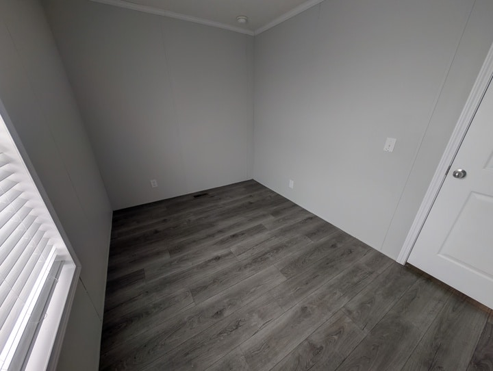 An empty room with light gray walls, dark wood-style flooring, a white door, several electrical outlets, and a window with blinds letting in natural light.