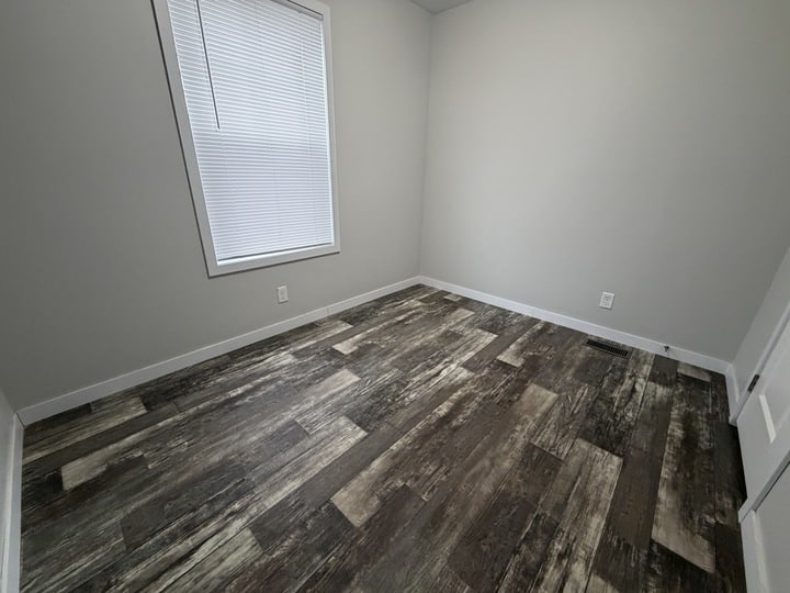 Empty room with gray and white walls, a single window covered by closed blinds, dark wood-patterned vinyl flooring, and white baseboards. No furniture is present.