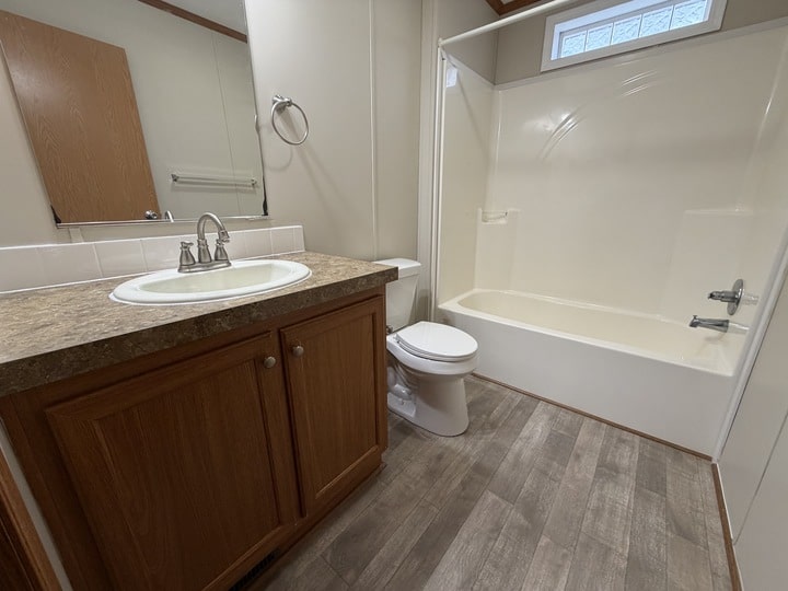 A clean bathroom with wood cabinets, a sink, toilet, and a bathtub-shower combo. The countertop is tan, floors are light wood, and walls are beige. A mirror and towel ring are mounted above the sink.