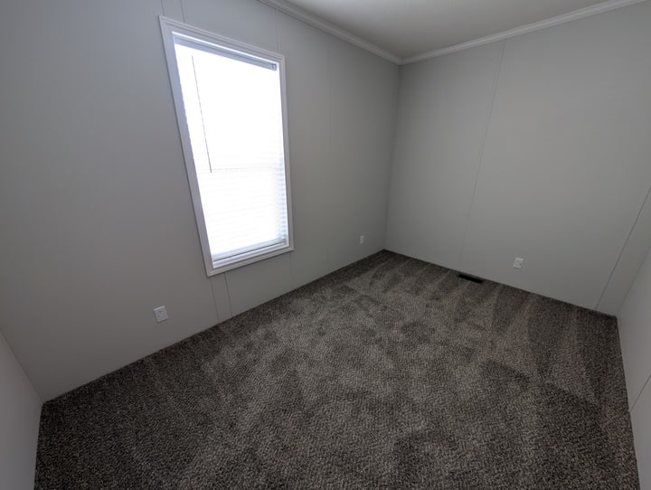 An empty room with light gray walls, a single window with blinds, and clean, textured gray carpet. There are two electrical outlets on each visible wall.