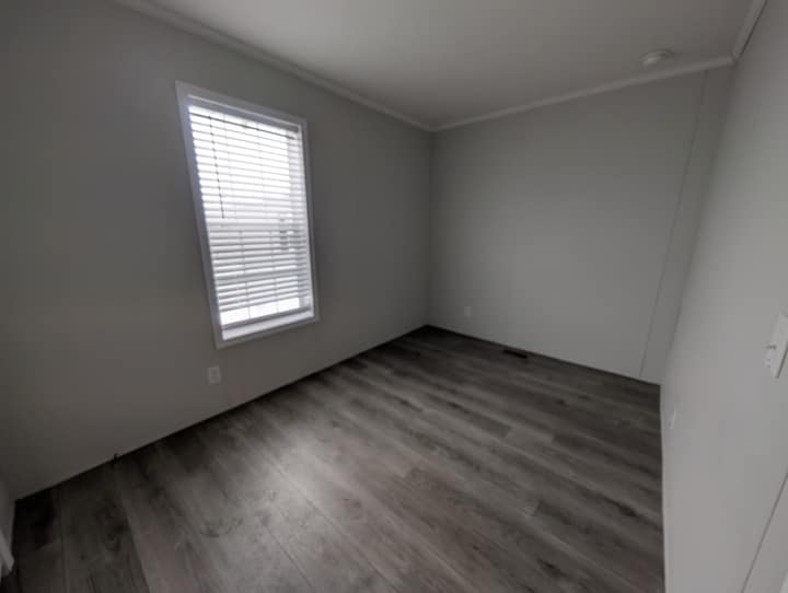 Empty room with light gray walls, a single window with white blinds, and gray wood-style flooring. The room appears clean and unfurnished, with natural light coming through the window.