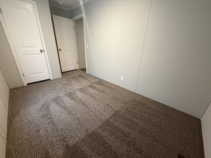 A small, empty room with light gray walls, beige carpet, and two white doors. The carpet shows fresh vacuum lines, and there is a white outlet on one wall. The room is well-lit and clean.