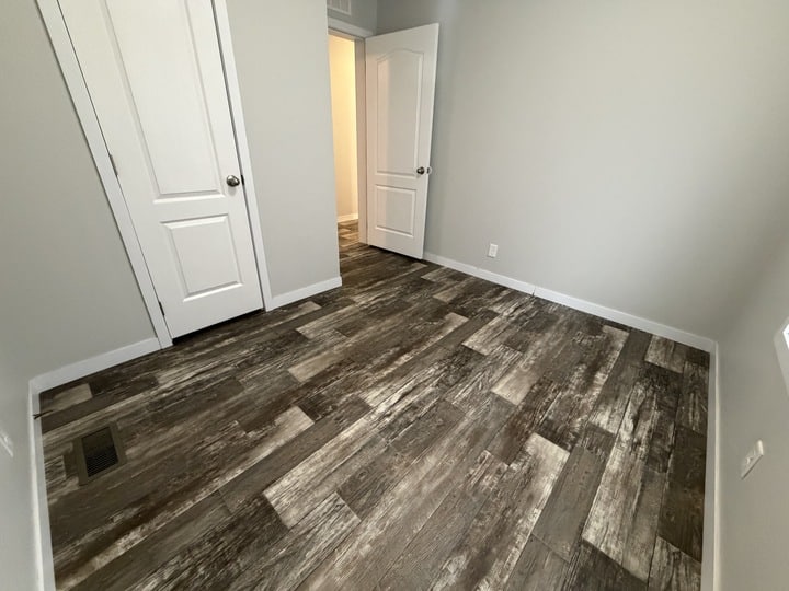 Empty room with grayish wood-look tile flooring, light gray walls, two white doors (one open, one closed), and a small vent on the floor near the wall. Warm light shines from the hallway.