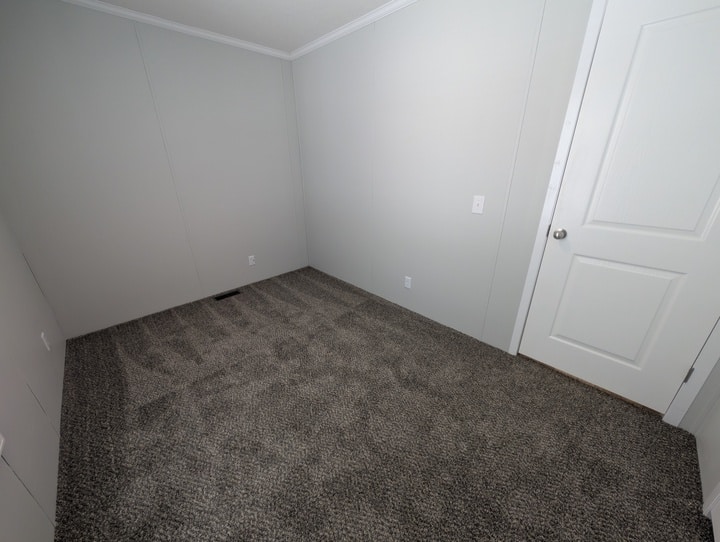 An empty room with light gray walls, white trim, a white door, and gray carpet. The room has no furniture or decorations and appears clean and newly finished.