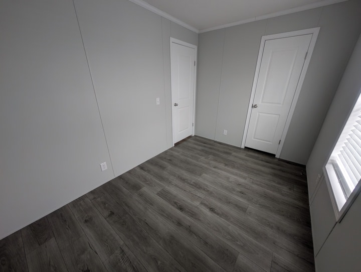 A small, empty room with light gray walls, dark wood-style flooring, two white doors, and a window with closed white blinds letting in natural light from the right side.