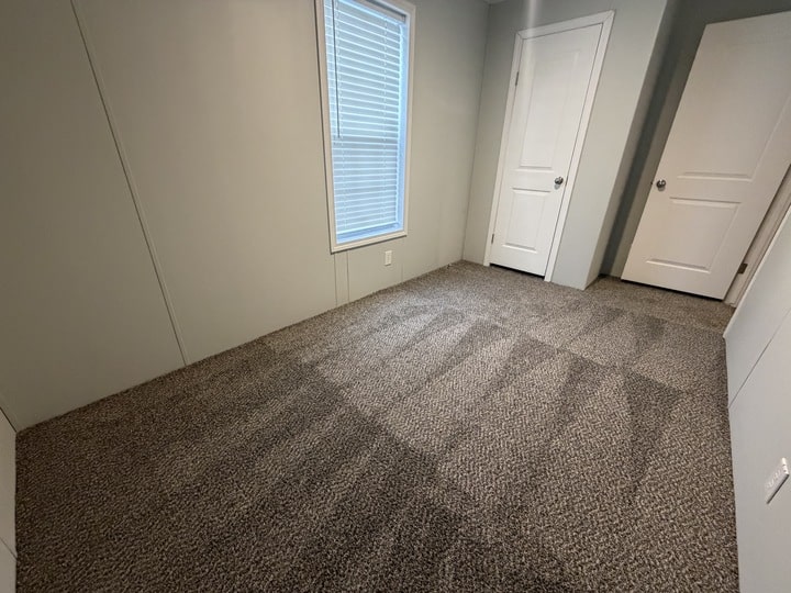 A small, empty room with light gray walls, a window with closed blinds, two white doors, and freshly vacuumed carpet.
