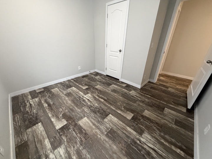 Small empty room with grayish wood-look vinyl flooring, light gray walls, white trim, and two white paneled doors—one closed, one open to a hallway with similar flooring. No furniture or decorations are present.