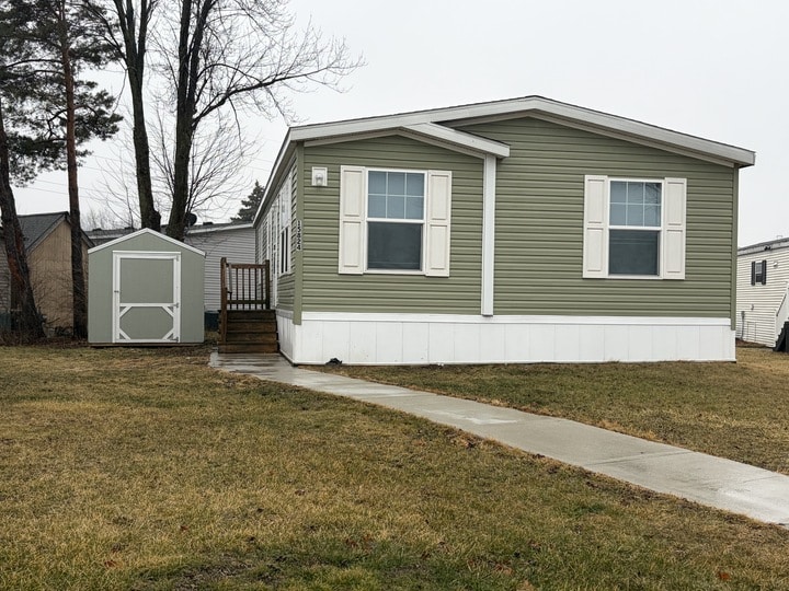 A green manufactured home with white trim and shutters, a small front porch with stairs, and a matching green shed beside it. The yard is grassy with a concrete walkway leading to the porch.