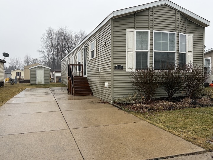 A beige manufactured home with white shutters, a small front porch, and a long concrete driveway on a cloudy day. Theres a shed in the backyard and bare bushes along the house. Grass and other homes are visible nearby.