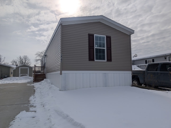 A beige mobile home with white trim and brown shutters is shown on a snowy lot under a cloudy sky. A pickup truck is parked beside the home, and there is a shed and driveway visible.
