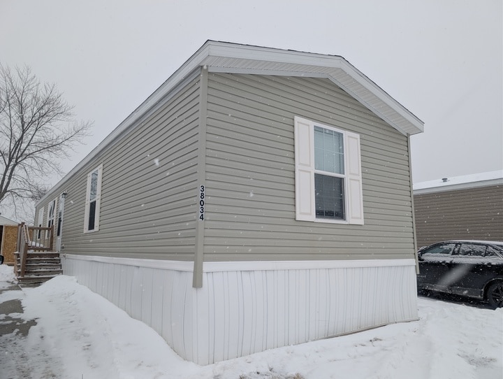 A light gray manufactured home with white trim and shutters is pictured on a snowy day. Snow covers the ground, and a leafless tree stands nearby. A black car is parked beside the home.