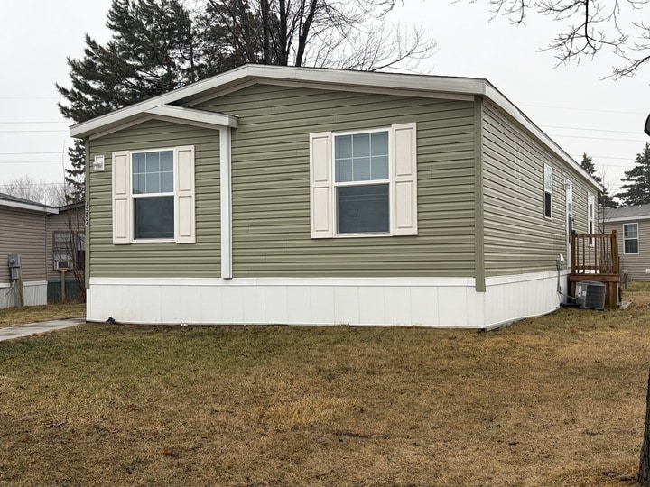 A green manufactured home with white trim, shutters, and skirting sits on a patchy, brown lawn. There are neighboring homes and bare trees in the background on an overcast day.