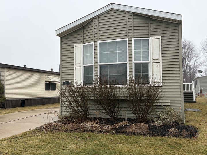 A light gray manufactured home with closed white shutters and large front windows, surrounded by leafless shrubs in a small garden bed, sits beside a paved driveway on a cloudy day. Another similar home is visible in the background.