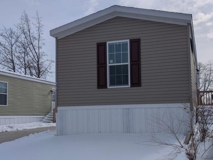 A beige mobile home with white trim and brown shutters sits on a snowy lot next to another similar home. Bare trees and a cloudy sky are visible in the background.