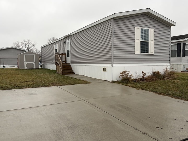 A gray mobile home with white trim sits on a concrete driveway, with a small wooden porch and steps leading to the entrance. There is a shed and patchy grass in the yard. The sky is overcast.