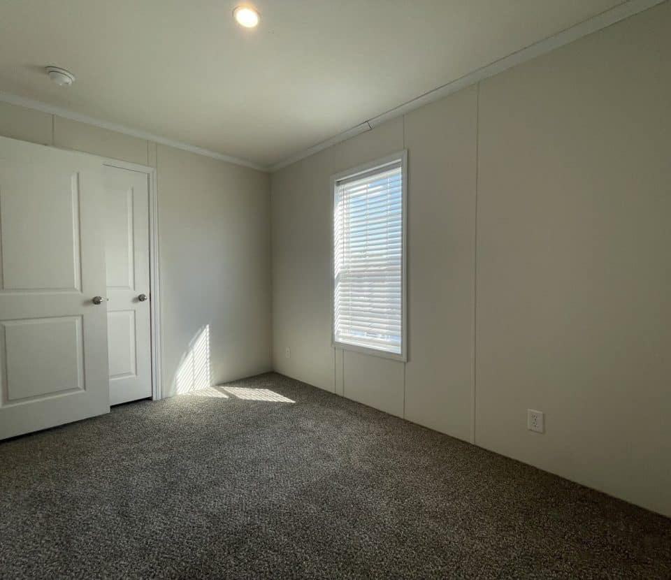 A small, empty room with beige walls, gray carpet, a closed window with blinds, and an open white door letting in sunlight. Ceiling light is on. The room appears clean and newly finished.